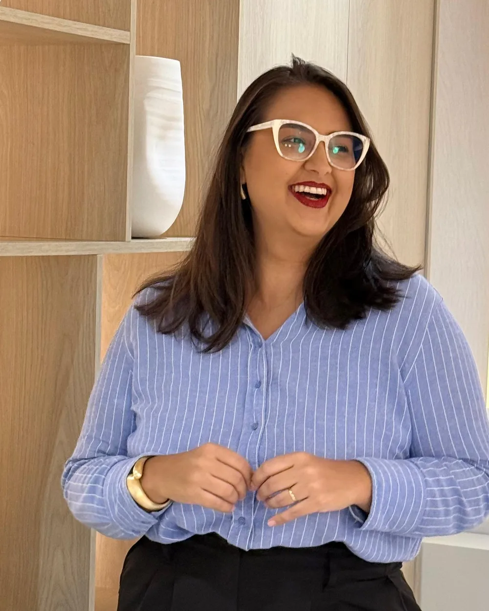 Cláudia Braz sorrindo, de camisa listrada azul, em frente a uma estante de madeira